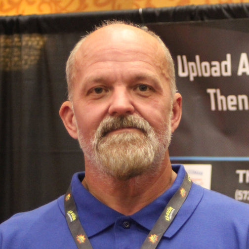 Man wearing a blue shirt with a lanyard, standing in front of a promotional backdrop.